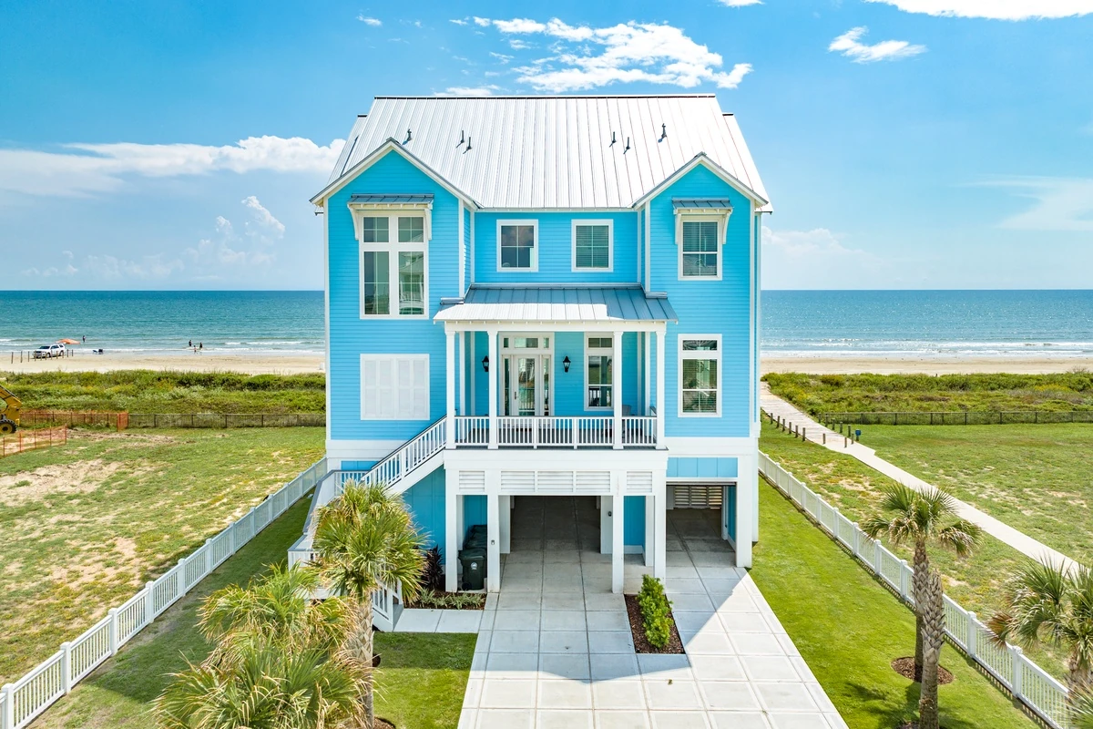 Bright turquoise elevated beach house with white trim, palm trees and a sunny blue sky beside the beach